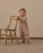 A baby standing on a patterned rug next to a rustic wooden chair, wearing a dusty pink long-sleeve jumpsuit with a subtle star pattern.