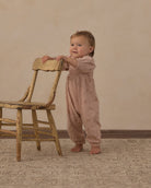A baby standing on a patterned rug and leaning on a rustic wooden chair, wearing a dusty pink smocked jumpsuit with a subtle celestial print.