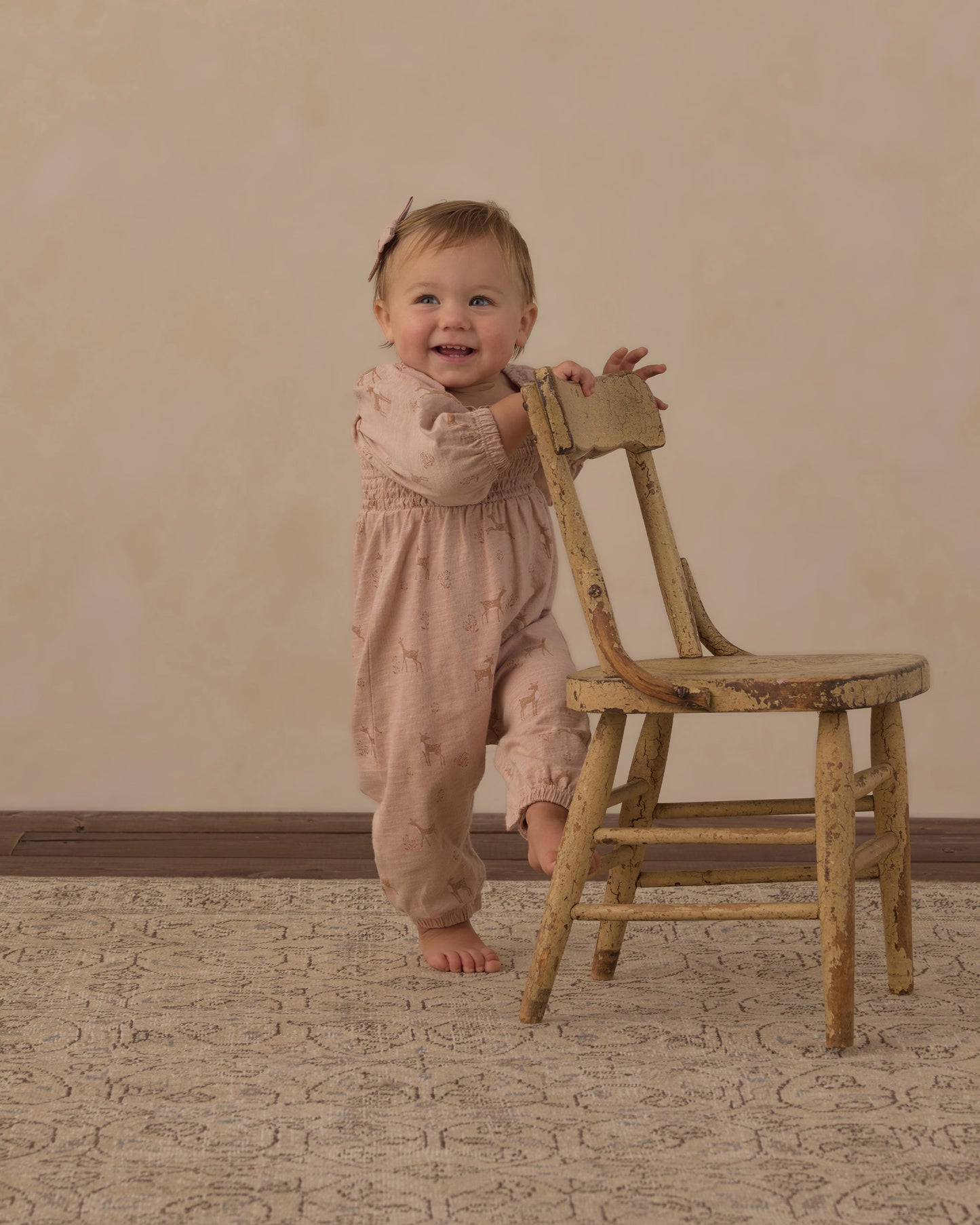 A baby standing barefoot next to a rustic wooden chair, wearing a dusty pink smocked jumpsuit with tiny embroidered details, set against a warm beige wall and patterned rug.