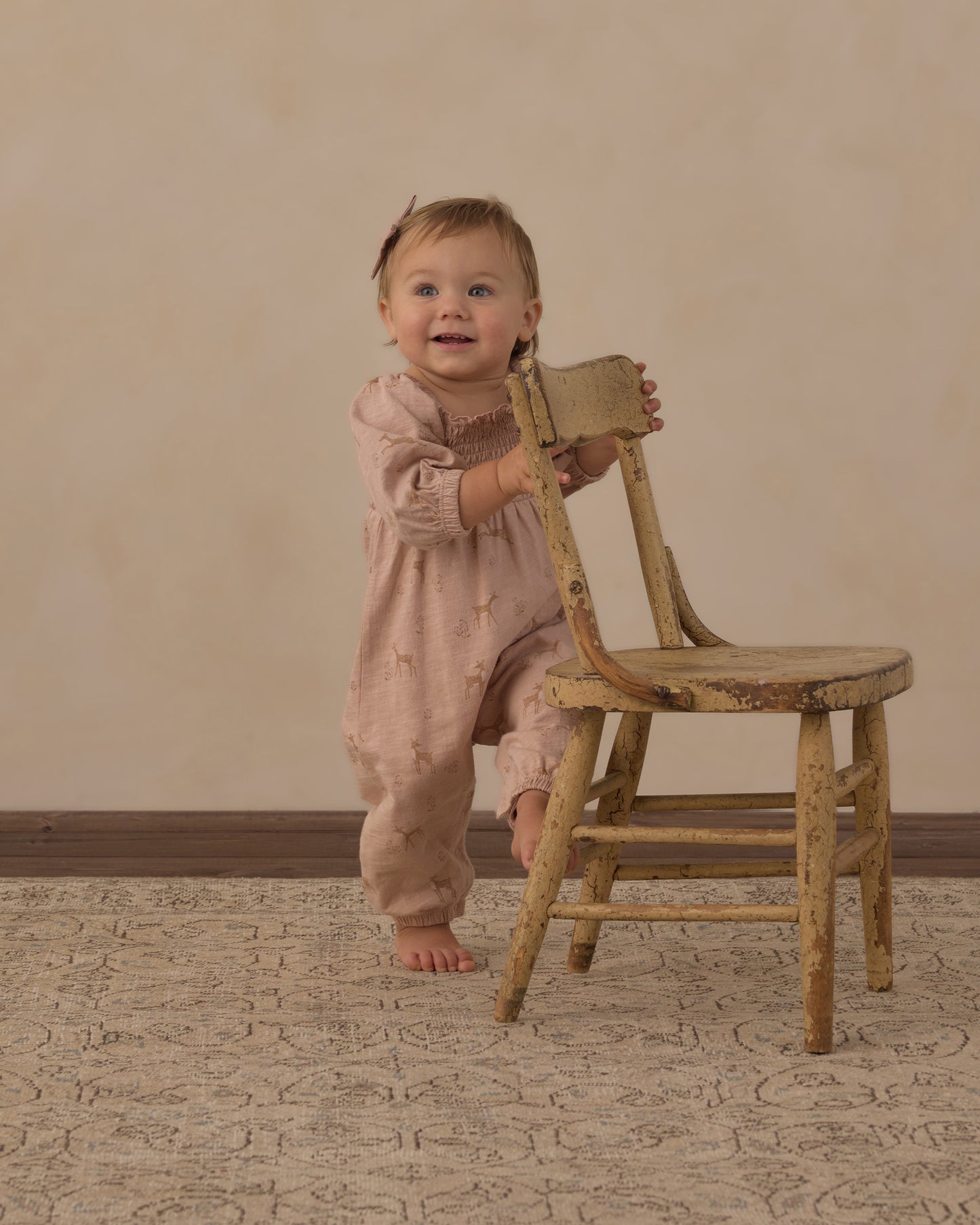 A baby wearing a dusty pink smocked jumpsuit stands next to a rustic wooden chair, smiling and looking at the camera against a warm beige studio backdrop.