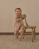 A baby wearing a dusty pink smocked jumpsuit stands next to a rustic wooden chair, smiling and looking at the camera against a warm beige studio backdrop.