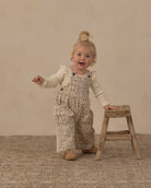 A toddler girl wearing beige and brown leopard-print overall with a cream long-sleeve shirt stands on a textured beige floor next to a small wooden stool, smiling and looking to the side.