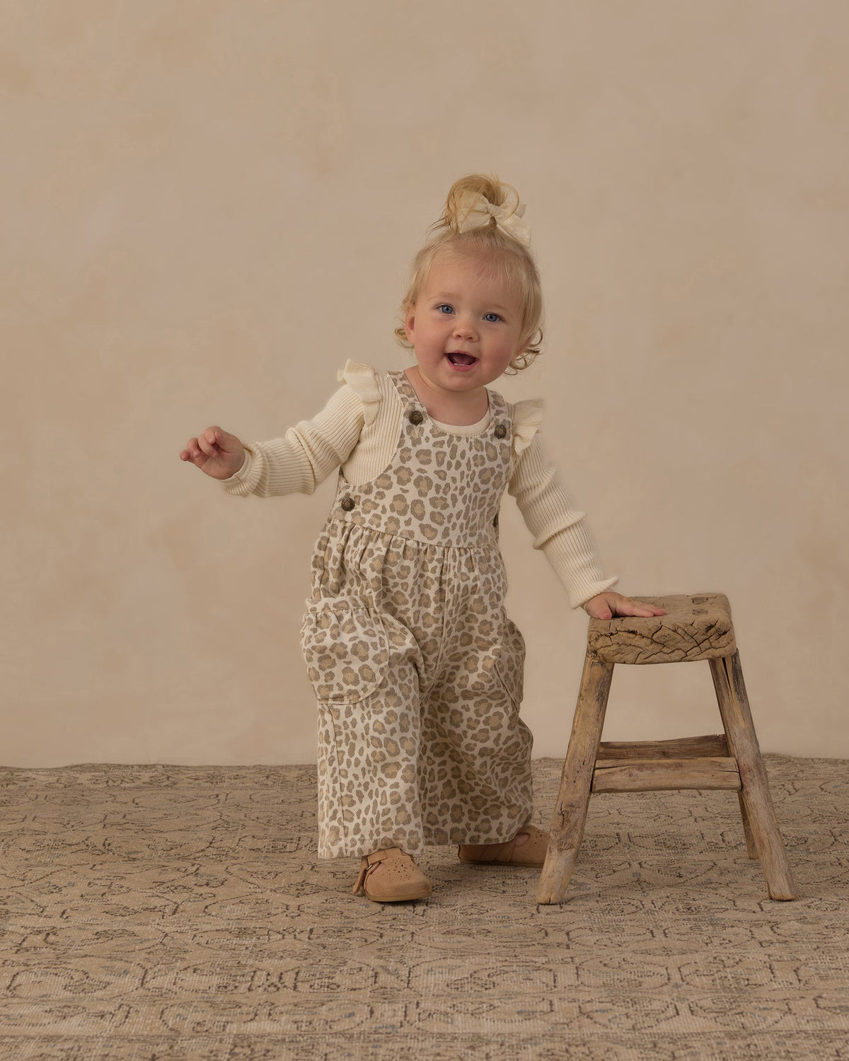 A toddler girl wearing beige and brown leopard-print overall with a cream long-sleeve shirt stands on a textured beige floor next to a small wooden stool, smiling and looking to the side.