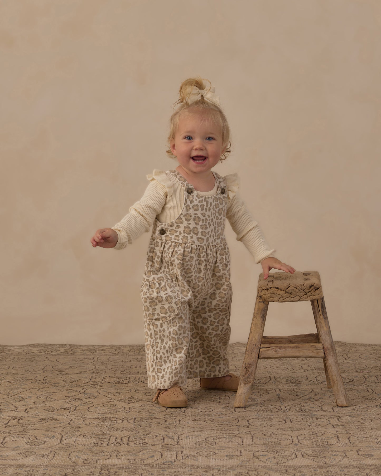  toddler girl wearing beige and brown leopard-print overall with a cream long-sleeve shirt stands on a textured beige floor next to a small wooden stool.