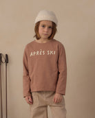 A child wearing a dusty rose long-sleeve sweatshirt with 'APRÈS SKI' printed in white across the chest, paired with beige and white vertically striped pants and a cream knit beanie, standing against a plain beige wall.