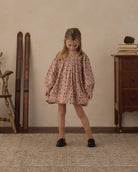 A young girl stands in a vintage-style room wearing a dusty rose pink smocked blouse with a small floral print, paired with beige socks and black shoes.