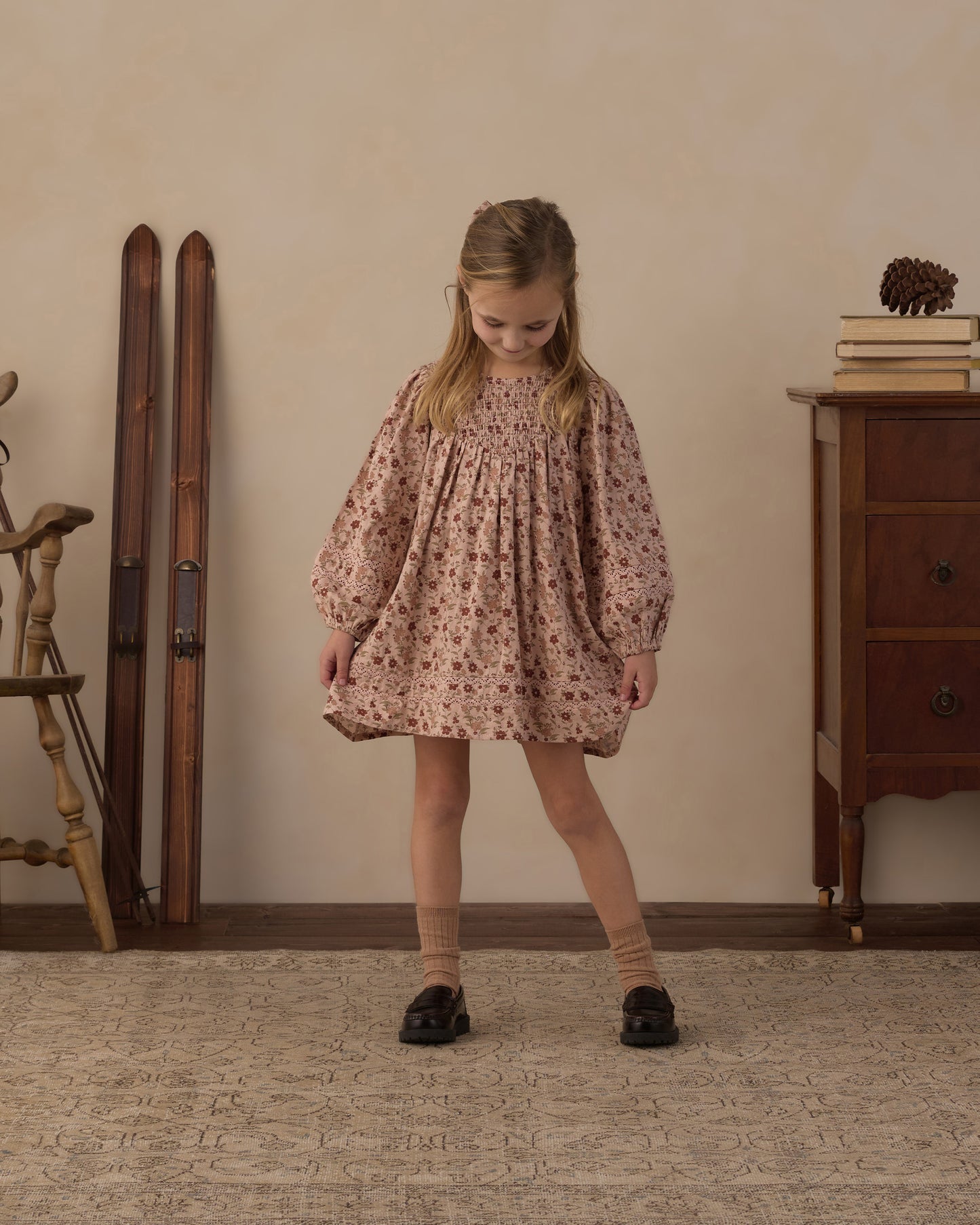 A young girl stands in a vintage-style room wearing a dusty rose pink smocked blouse with a small floral print, paired with beige socks and black shoes.