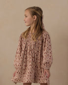 A young girl with blonde hair tied back is wearing a long-sleeved, smocked babydoll dress with a dusty rose and brown floral pattern, photographed against a neutral beige background.