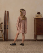 A young girl is shown from behind wearing a dusty pink floral dress with a matching large bow in her hair, standing in a vintage-style room with a wooden dresser, books, and skis in the background.