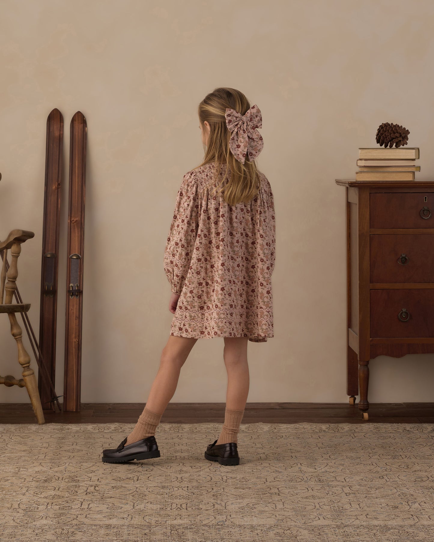 A young girl is shown from behind wearing a dusty pink floral dress with a matching large bow in her hair, standing in a vintage-style room with a wooden dresser, books, and skis in the background.