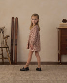 A young girl with blonde hair stands in a rustic room wearing a long-sleeved, knee-length pink dress with a small floral pattern, paired with beige socks and black loafers.