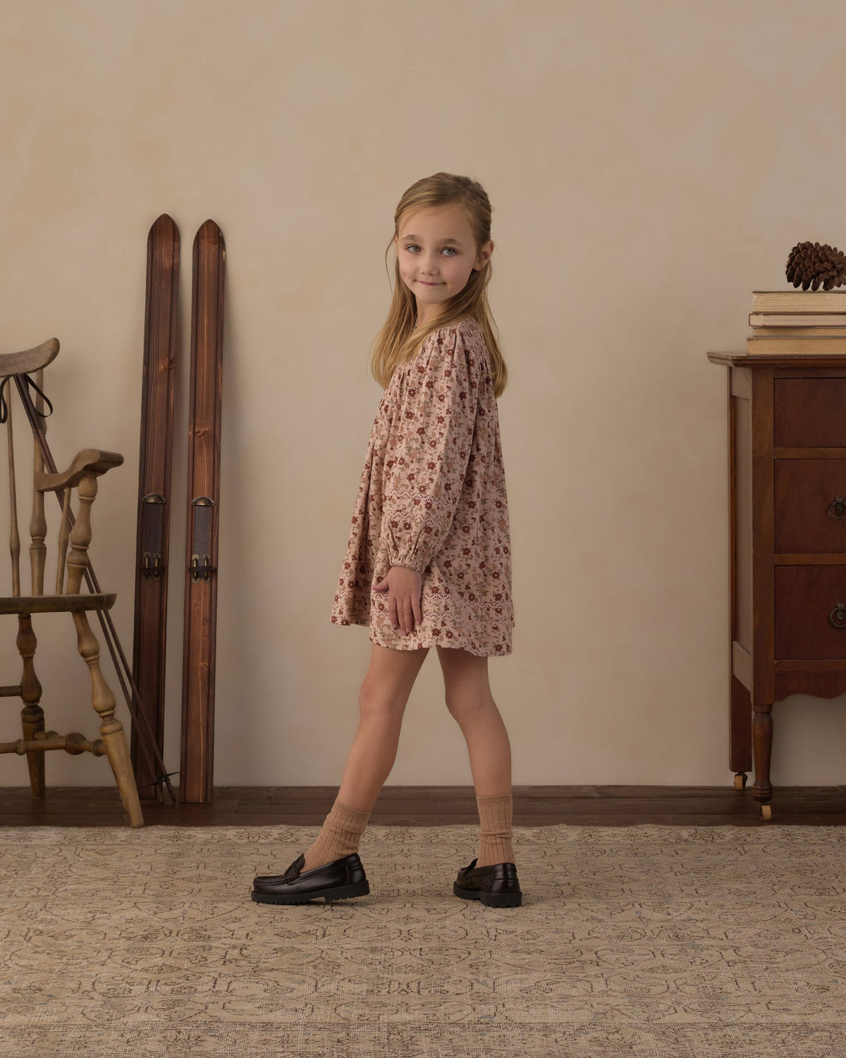A young girl with blonde hair stands in a rustic room wearing a long-sleeved, knee-length pink dress with a small floral pattern, paired with beige socks and black loafers.