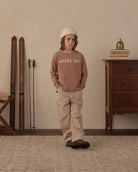 A child stands in a styled indoor room wearing a dusty rose long-sleeve sweatshirt with 'APRÈS SKI' printed in white across the chest, paired with beige corduroy pants and a white knit beanie. Ski boots and skis lean against the wall on the left, and a wooden cabinet with books and a small brass bell sits on the right.