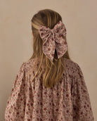 A blonde girl viewed from behind, wearing a large dusty pink floral hair bow accessory in her hair, paired with a matching smocked blouse.