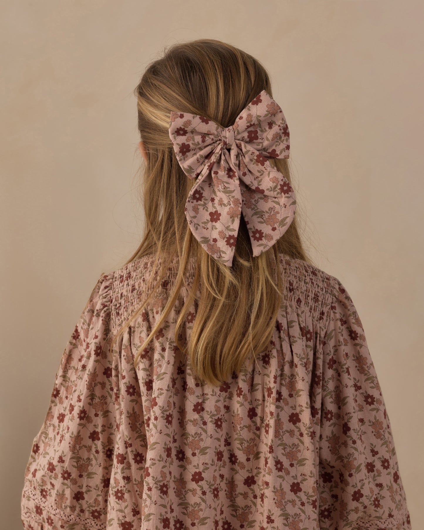 A blonde girl viewed from behind, wearing a large dusty pink floral hair bow accessory in her hair, paired with a matching smocked blouse.