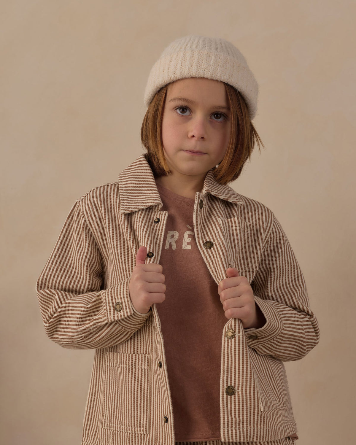 A young child wearing a cream knit beanie, a brown graphic t-shirt, and a beige and brown vertically striped button-up overshirt, posing against a solid beige background.