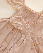 A close-up of a baby dress in a dusty pink color, featuring a smocked bodice, ruffled trim, and a printed pattern of small deer and floral motifs.