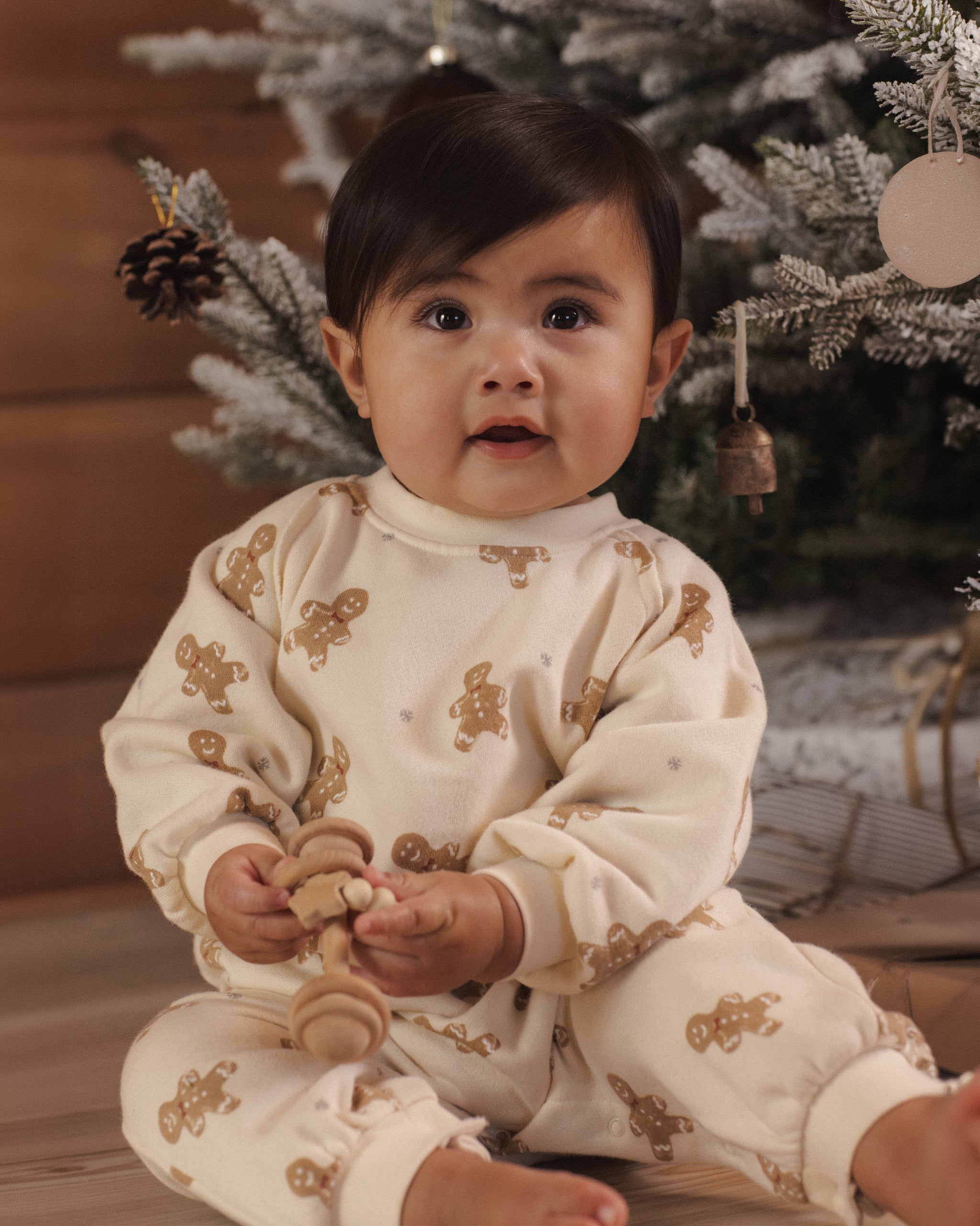 A baby sitting in front of a decorated Christmas tree, wearing a cream-colored long-sleeve pajama set with brown gingerbread cookie prints.