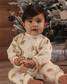 A baby sitting in front of a decorated Christmas tree, wearing a cream-colored long-sleeve pajama set with brown gingerbread cookie prints.