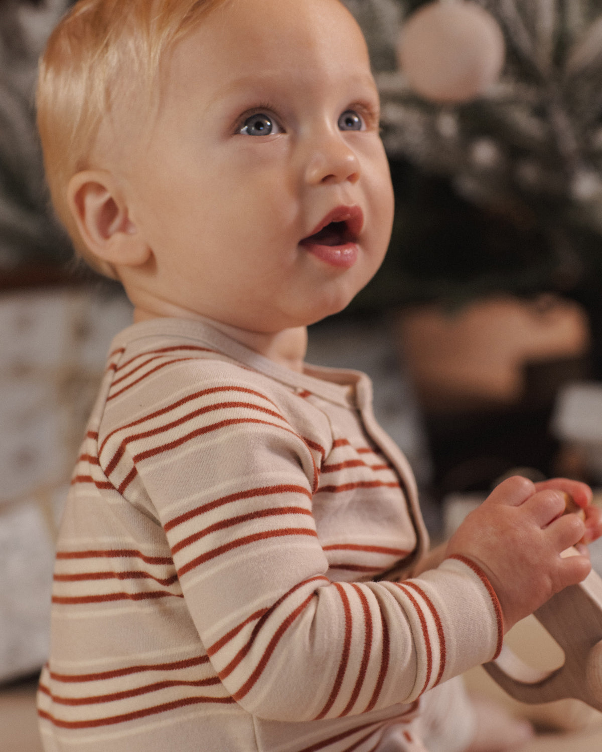 A close-up of a baby wearing a cream-colored long-sleeve romper with thin rust-brown horizontal stripes, looking off-camera with an open mouth.