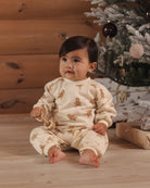 A baby sitting on a wooden floor in front of a Christmas tree, wearing a cream-colored one-piece pajama with a brown gingerbread man pattern.