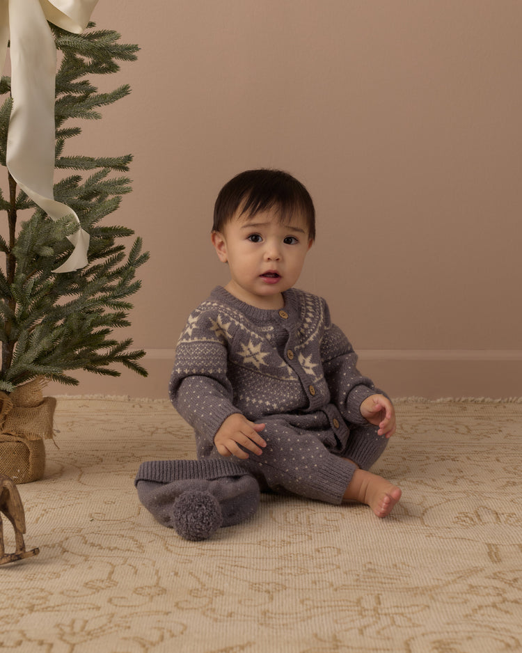A baby sitting on a patterned rug next to a decorated Christmas tree, wearing a gray knit cardigan and matching knit pants with cream-colored snowflake patterns.