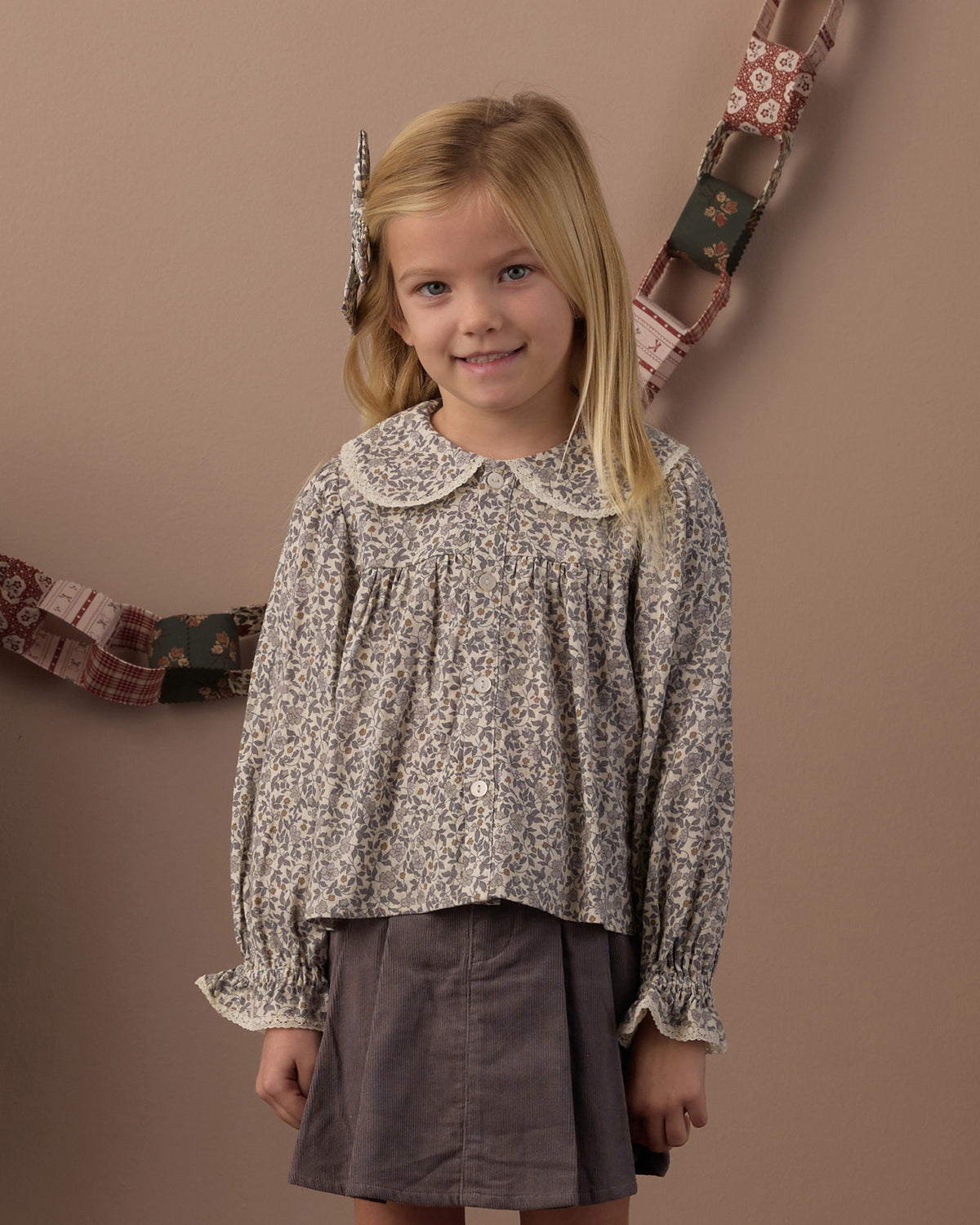 A young girl with blonde hair wearing a long-sleeved, vintage-style floral blouse with a Peter Pan collar and a dark gray skirt, standing against a warm brown backdrop with a decorative patchwork garland.