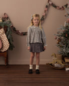 A young girl stands in a decorated indoor room wearing a long-sleeve patterned blouse with a Peter Pan collar and a dark gray skirt. The background features a warm beige wall with a garland, stockings, and a Christmas tree.