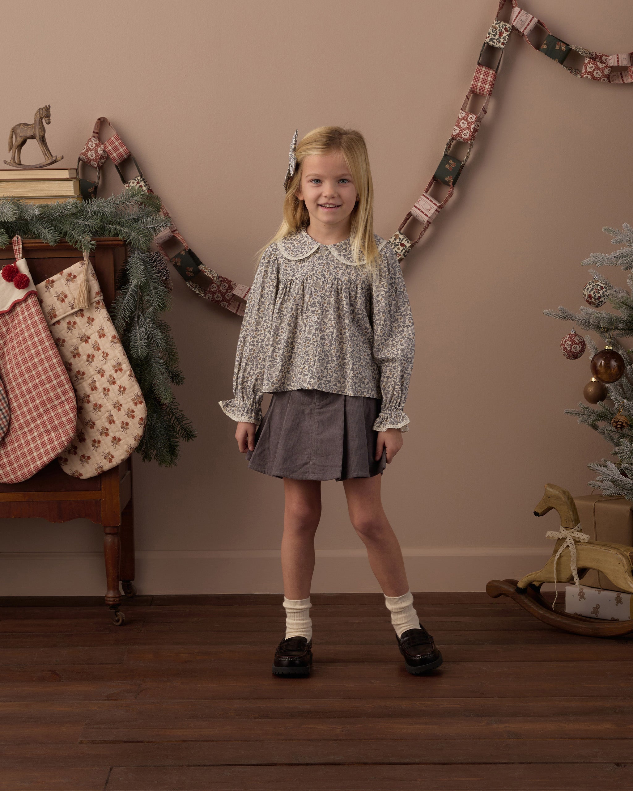 A young girl stands in a decorated indoor room wearing a long-sleeve patterned blouse with a Peter Pan collar and a dark gray skirt. The background features a warm beige wall with a garland, stockings, and a Christmas tree.