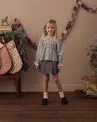 A young girl stands in a decorated indoor room wearing a long-sleeve patterned blouse with a Peter Pan collar and a dark gray skirt. The background features a warm beige wall with a garland, stockings, and a Christmas tree.