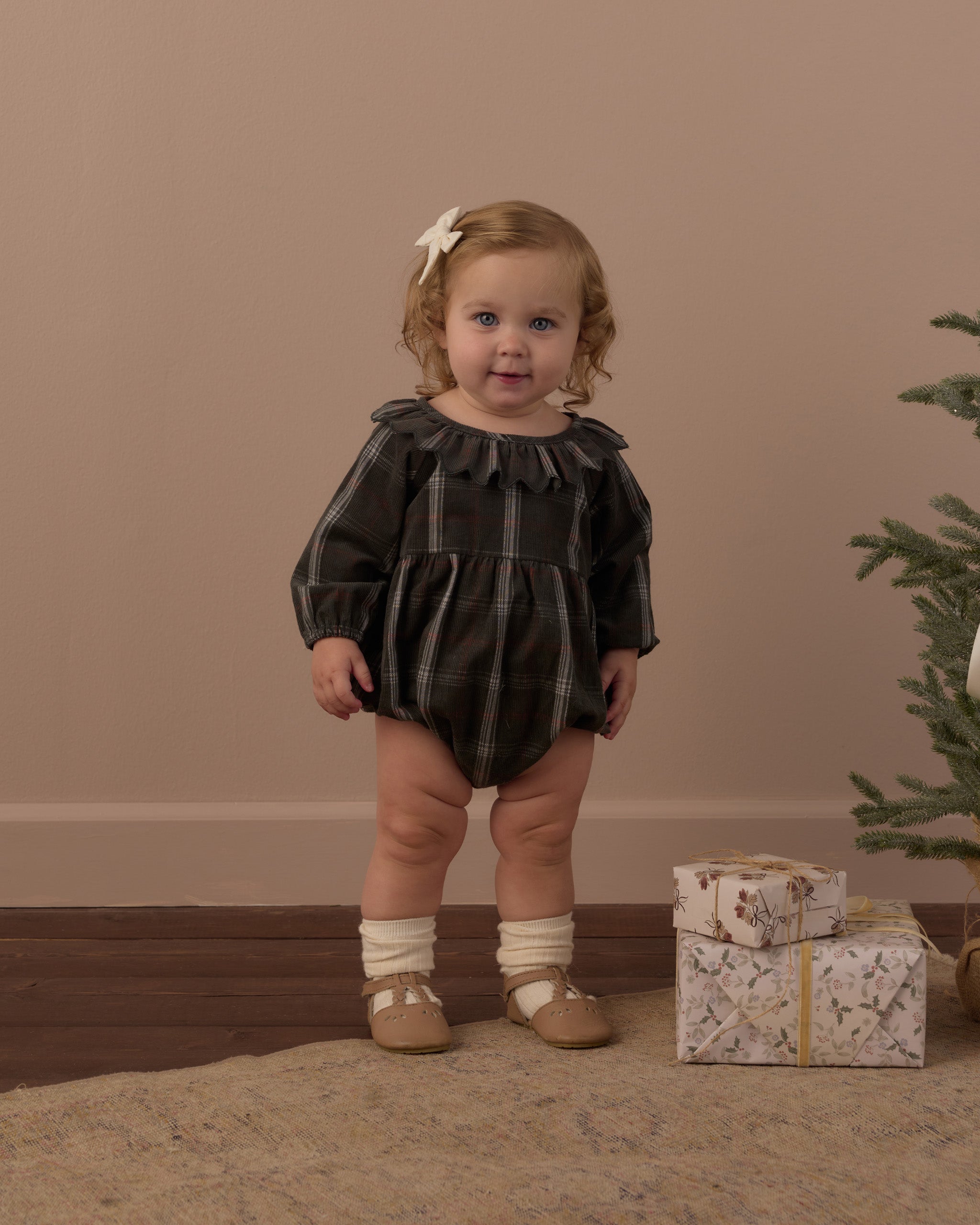 A toddler stands indoors wearing a dark plaid romper with long puff sleeves, cream socks, and tan shoes. The room has a warm beige wall, wood floor, a small Christmas tree, and wrapped gifts on the floor.