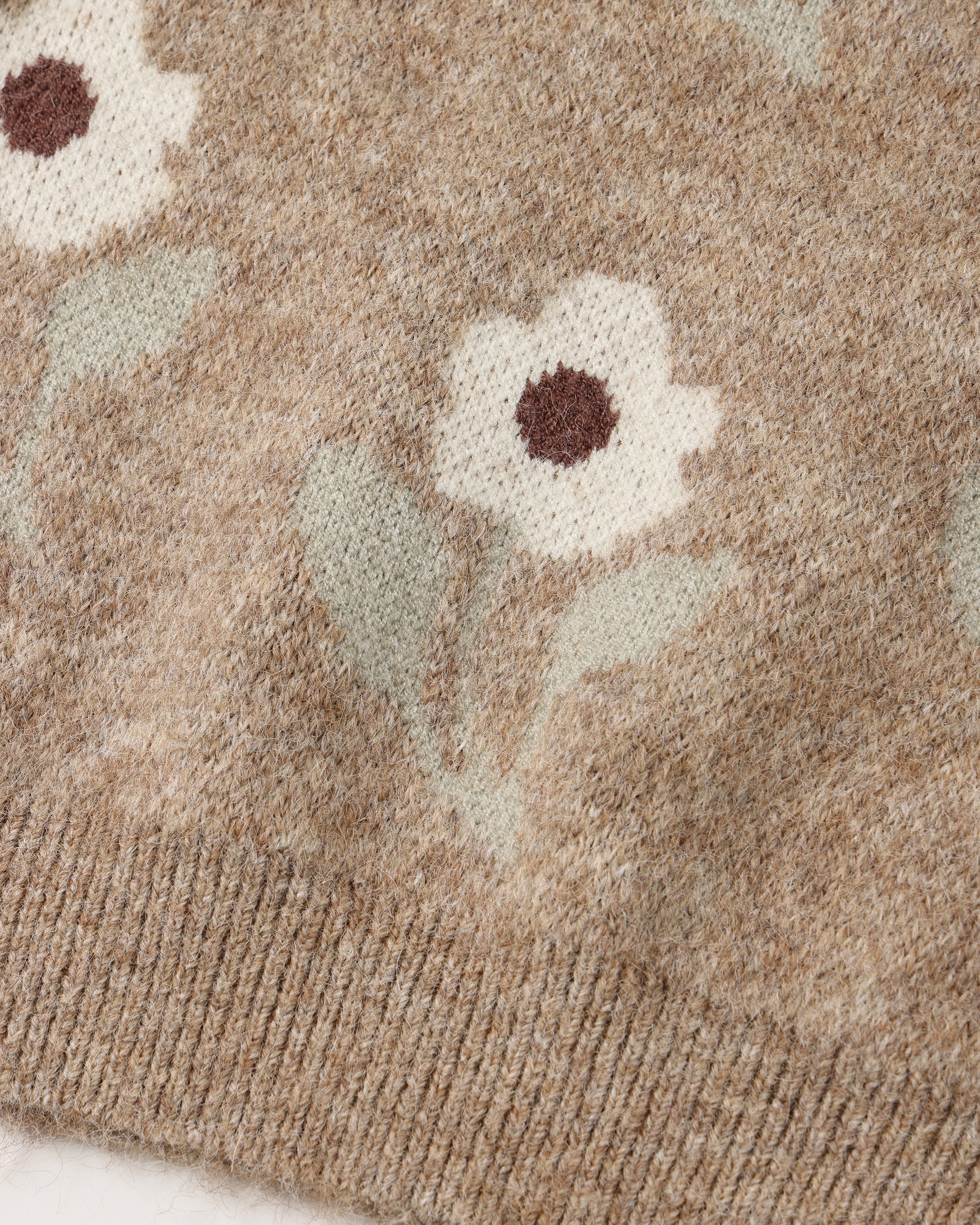 Close-up of a knit sweater showing a floral pattern in cream, sage green, and brown on a heathered taupe-beige background.