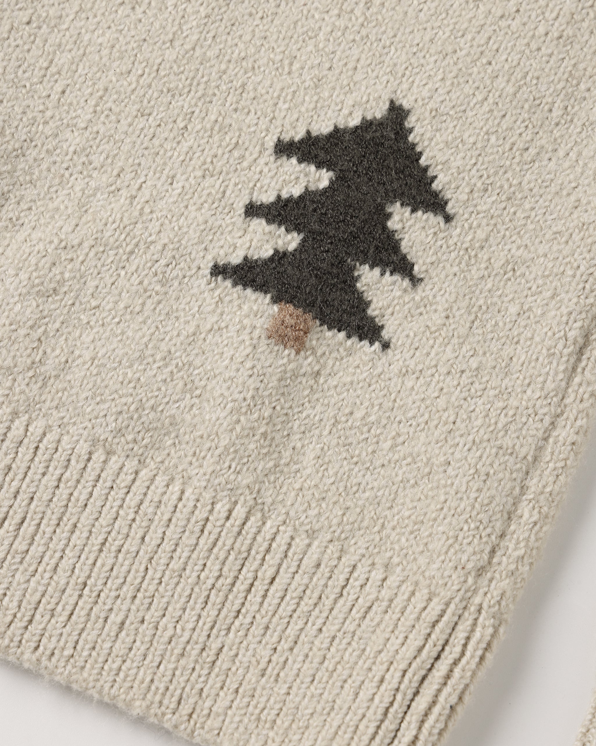 A close-up view of a cream-colored knit sweater featuring a dark green pine tree pattern with a brown trunk and ribbed texture at the hem.