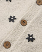 Close-up of a cream-colored textured knit fabric featuring embroidered dark gray flower motifs and natural brown wooden buttons.