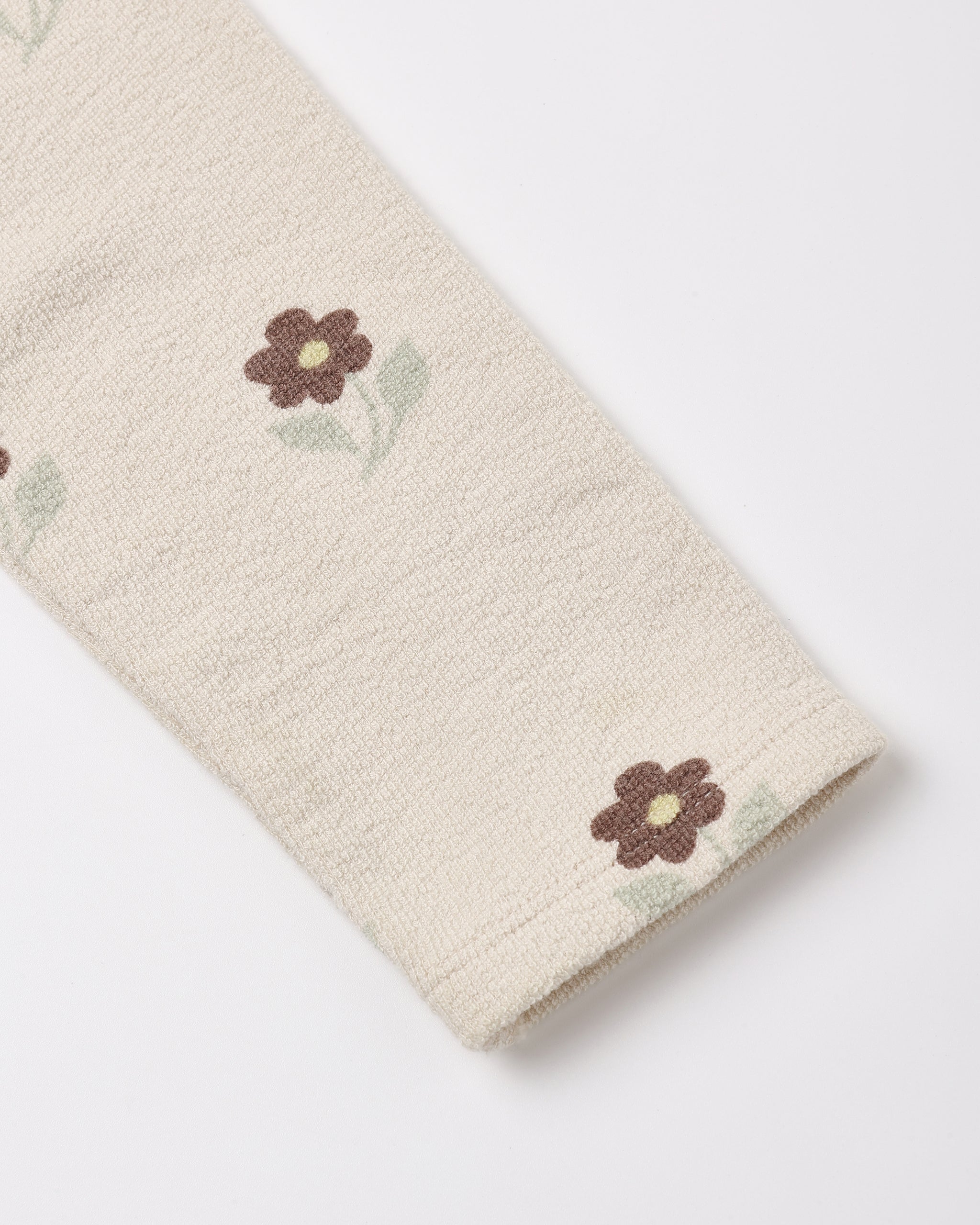Close-up of a cream-colored textured fabric, likely a sweater or knit garment, featuring a small brown and green embroidered flower pattern.