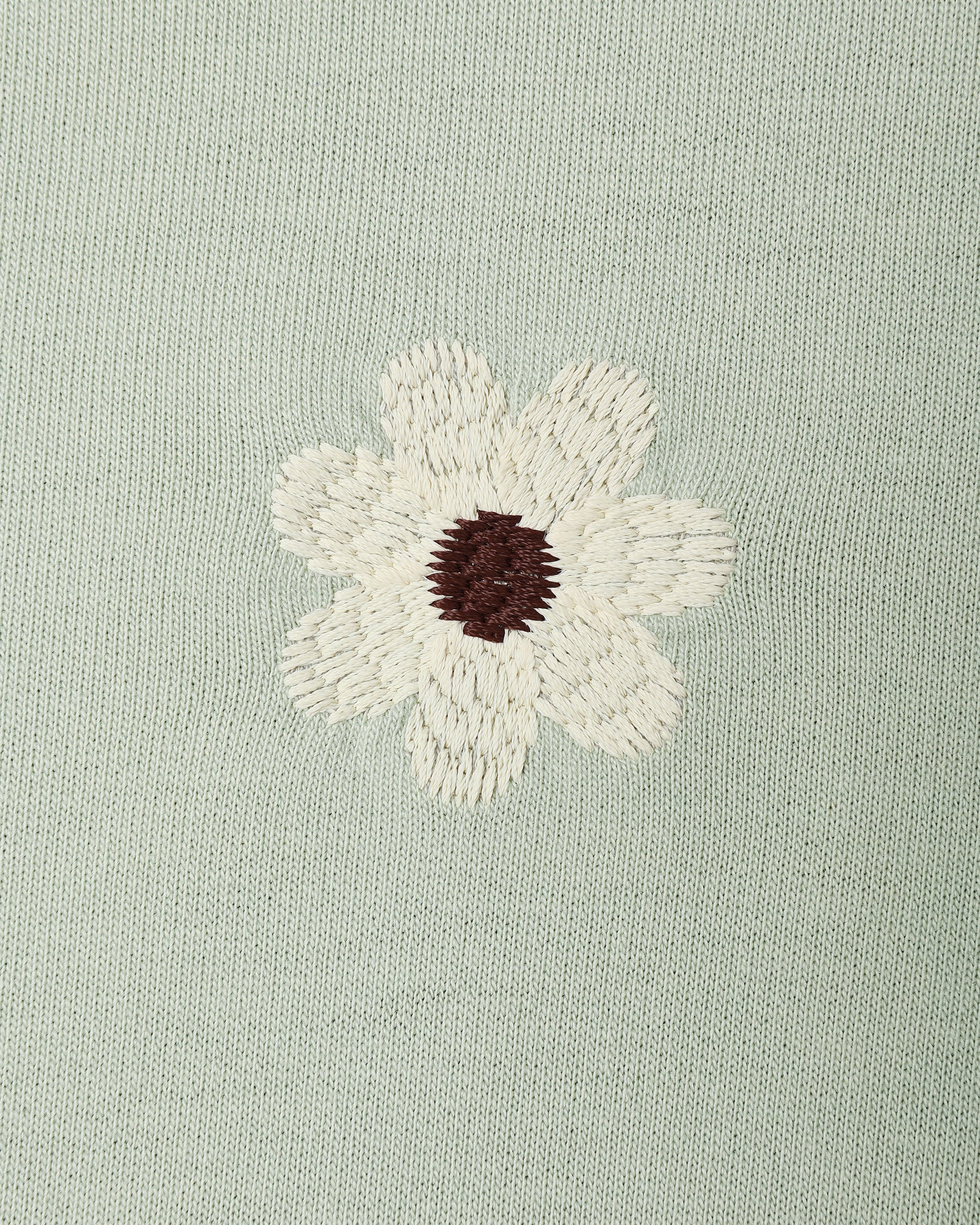 A close-up of a light sage green knit fabric featuring an embroidered daisy flower with cream petals and a dark brown center.