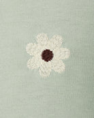 A close-up of a light sage green knit fabric featuring an embroidered daisy flower with cream petals and a dark brown center.