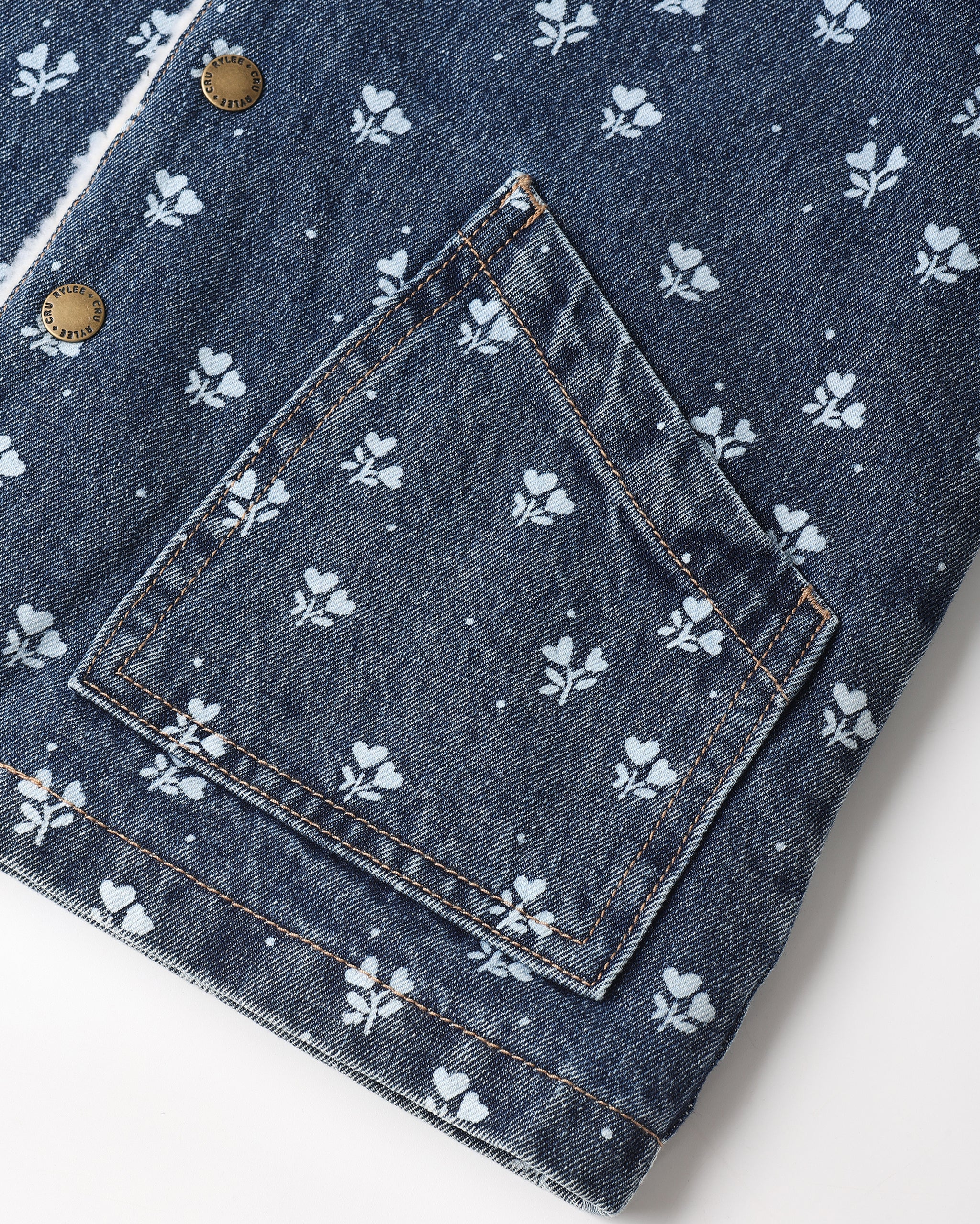 Close-up of a dark blue denim jacket featuring a white floral and dot pattern, a large patch pocket, and brass snap buttons.