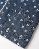 Close-up of a dark blue denim jacket featuring a white floral and dot pattern, a large patch pocket, and brass snap buttons.