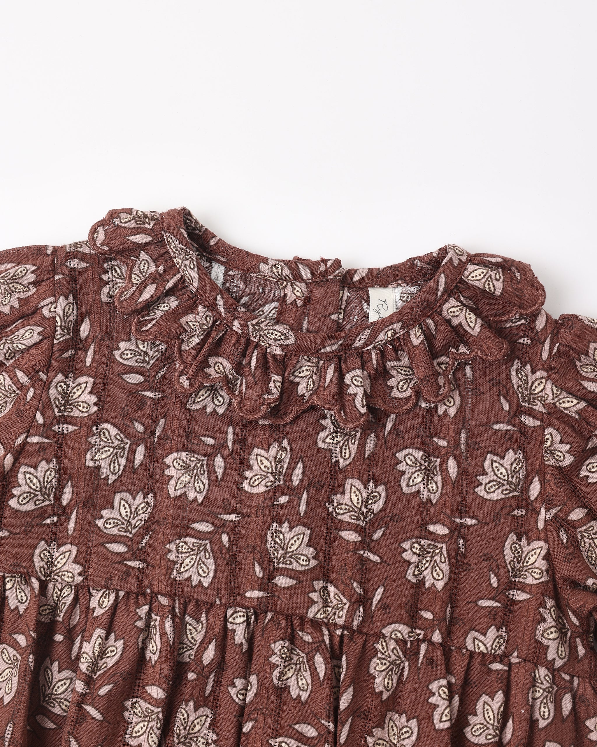 A close-up of a brown cotton dress with a white floral and leaf pattern, featuring a ruffled collar and tiered skirt.