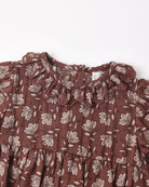 A close-up of a brown cotton dress with a white floral and leaf pattern, featuring a ruffled collar and tiered skirt.