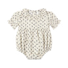 A baby romper in a warm off-white fabric with a small, repeating dark floral print, featuring short flutter sleeves and a ruffled neckline.