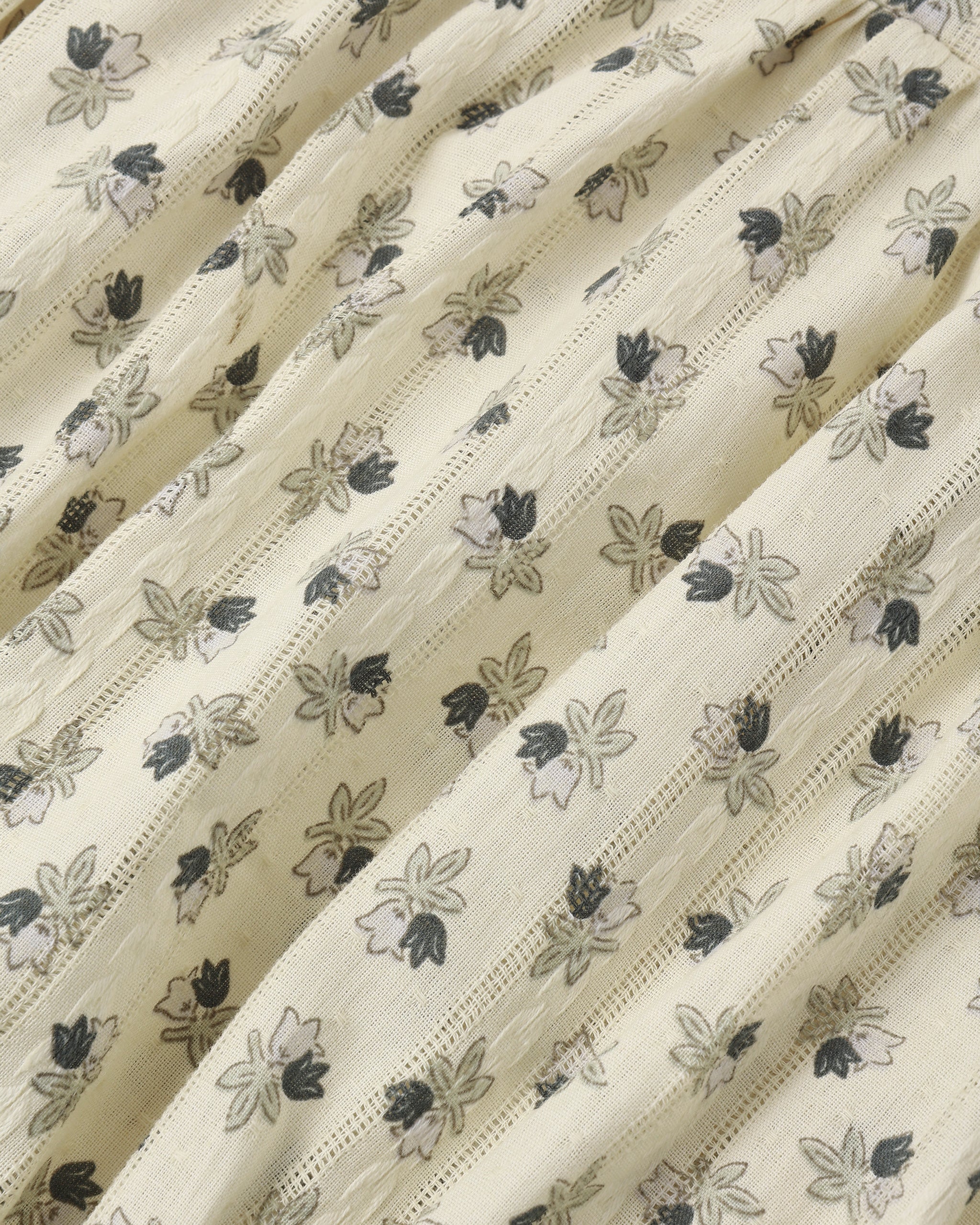 A close-up of a cream-colored fabric with a delicate, repeating floral block print in black and gray, featuring fine vertical stitched stripes.