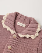 Close-up of a knitted cardigan in dusty mauve pink with a cream scalloped collar and light buttons, showing a brand label that reads 'Ryōn's Cream Sweaters'.