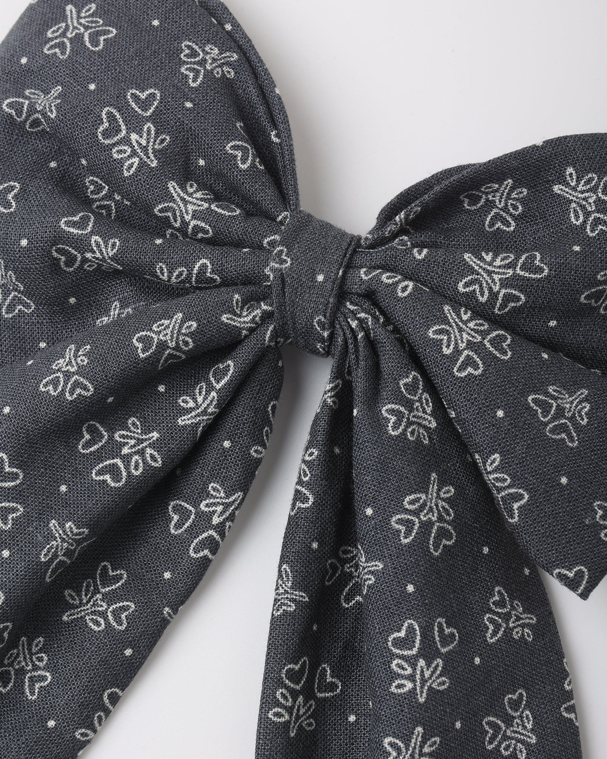 A close-up of a dark gray fabric bow with a white line-art pattern of small hearts and floral motifs, set against a plain light gray background.