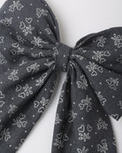 A close-up of a dark gray fabric bow with a white line-art pattern of small hearts and floral motifs, set against a plain light gray background.