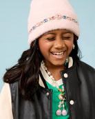 A young girl smiling while wearing a light pink fuzzy beanie with a jeweled headband, matching pink and blue teardrop earrings, and a pink and blue rhinestone necklace and wearing black jacket against a light blue background.
