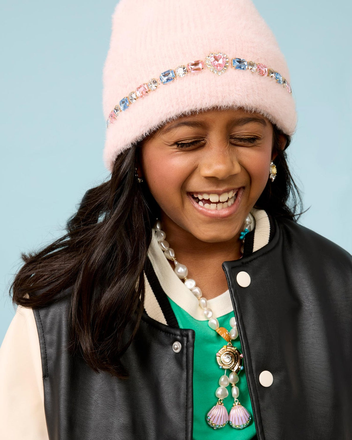 A young girl smiling while wearing a light pink fuzzy beanie with a jeweled headband, matching pink and blue teardrop earrings, and a pink and blue rhinestone necklace and wearing black jacket against a light blue background.