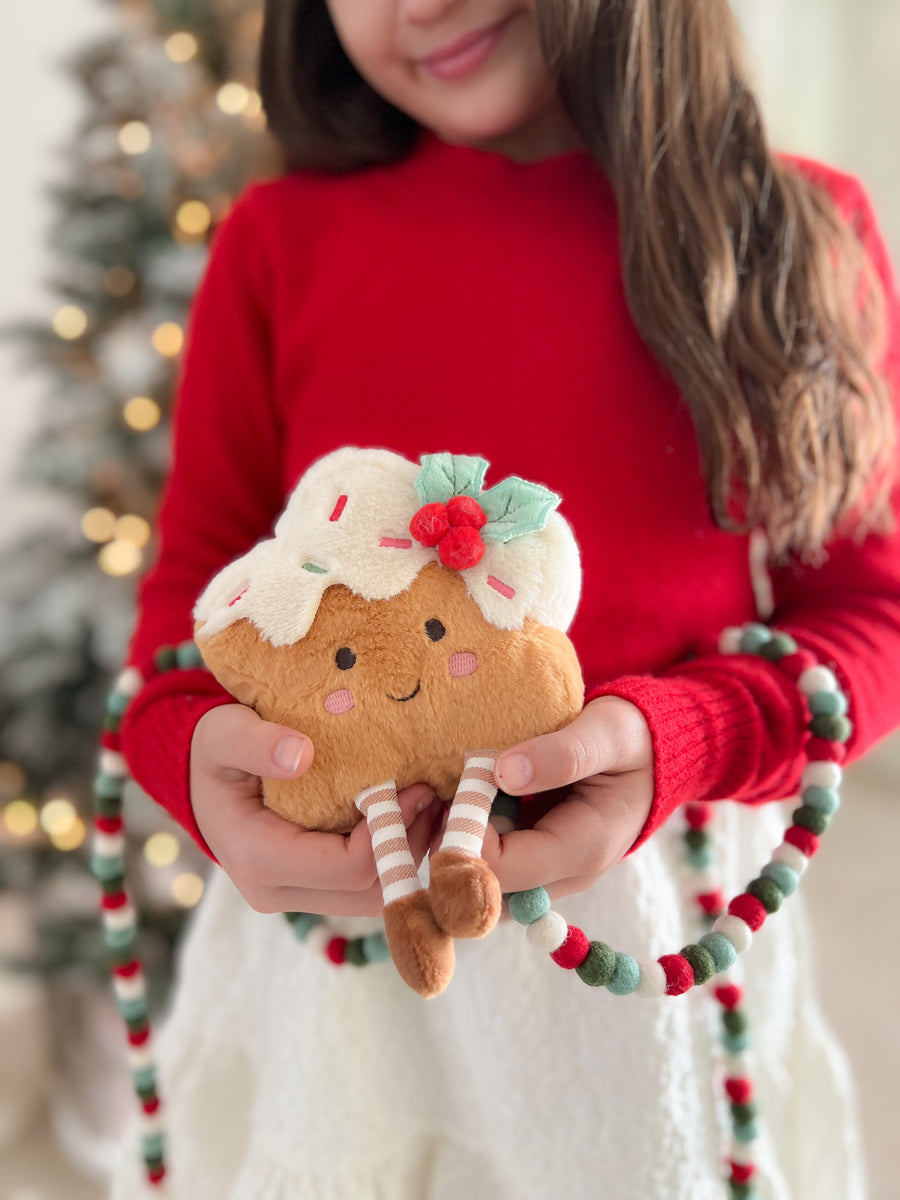 A child in a red sweater holds a cute plush cupcake toy with white frosting, a red cherry, and striped legs. The background shows a softly blurred Christmas tree with warm lights.
