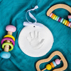 White ceramic handprint keepsake ornament with a white ribbon, surrounded by wooden baby toys on a teal background.